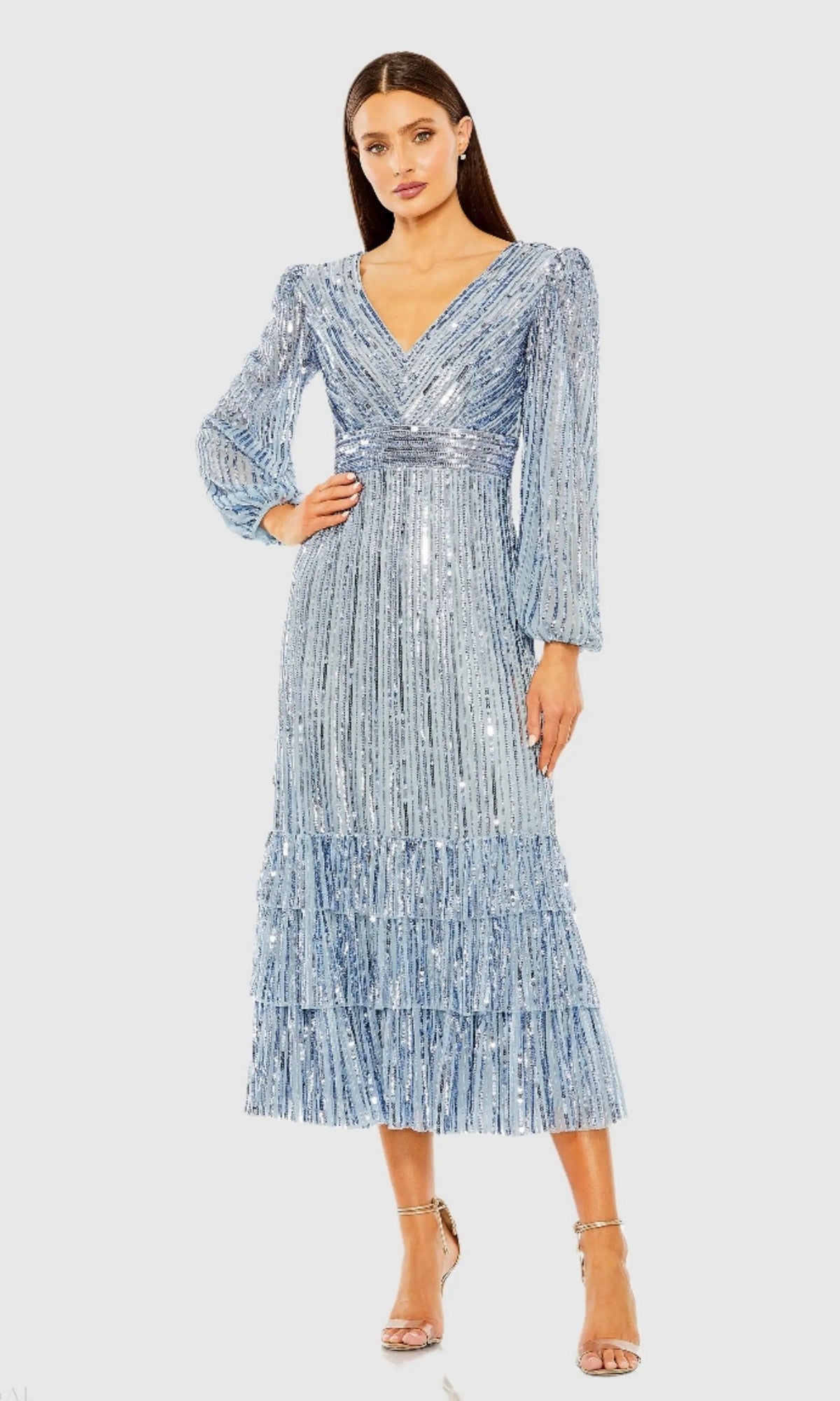 A woman wears the Mac Duggal 23006 Midi Semi-Formal Dress in slate blue, featuring sequins, long sleeves, a v-neckline, tiered skirt, and silver high-heeled sandals.